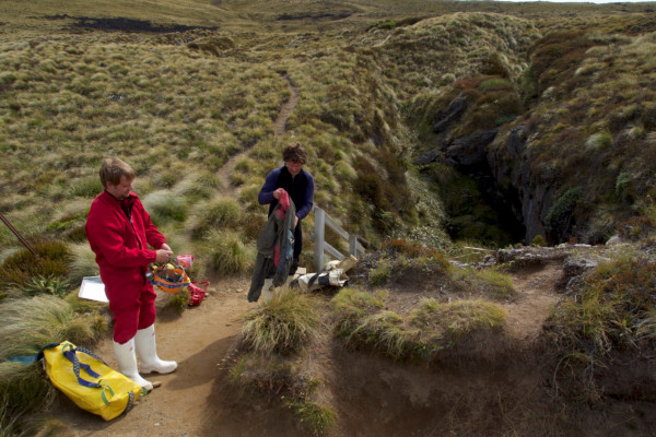 Two people put on caving equipment in fromnt of a cave entrance