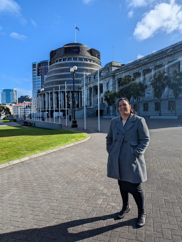 Dr Pauline Harris, in a winter coat, stands in front of the Beehive and houses of Parliament