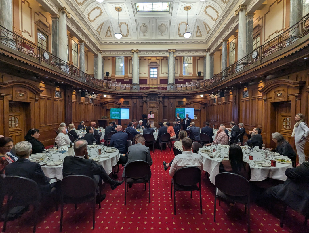 Long view of an ornate room with red carpet. Many MPs sit at dinner tables and the speakers stand in front of screens gesticulating.