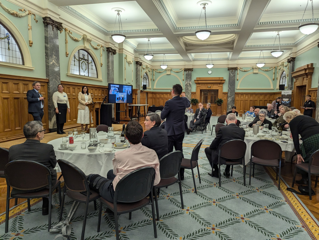 Prof Richard Easther, Dr Pauline Harris, and host Dr Parmjeet Parmar MP listen to a question from a male MP, who stands at a round table, amid seated guests, with his back to the camera